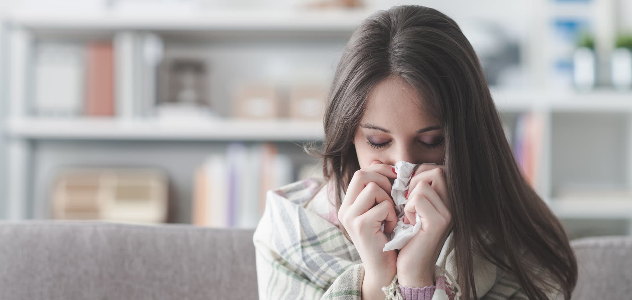 Sick young woman at home on the sofa with a cold, she is covering with a blanket and blowing her nose
